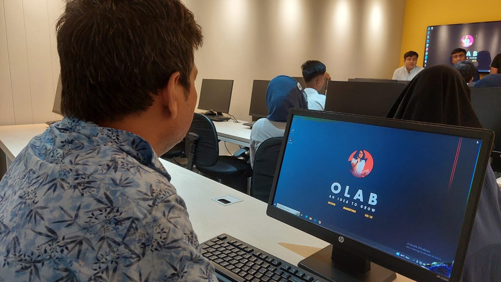 A learner at a computer in an OLab-branded classroom, with the instructor and other students in the background